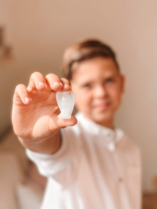 Een persoon houdt een klein beschermengeltje van bergkristal vast tussen duim en wijsvinger. De achtergrond is onscherp en de persoon draagt een wit overhemd.