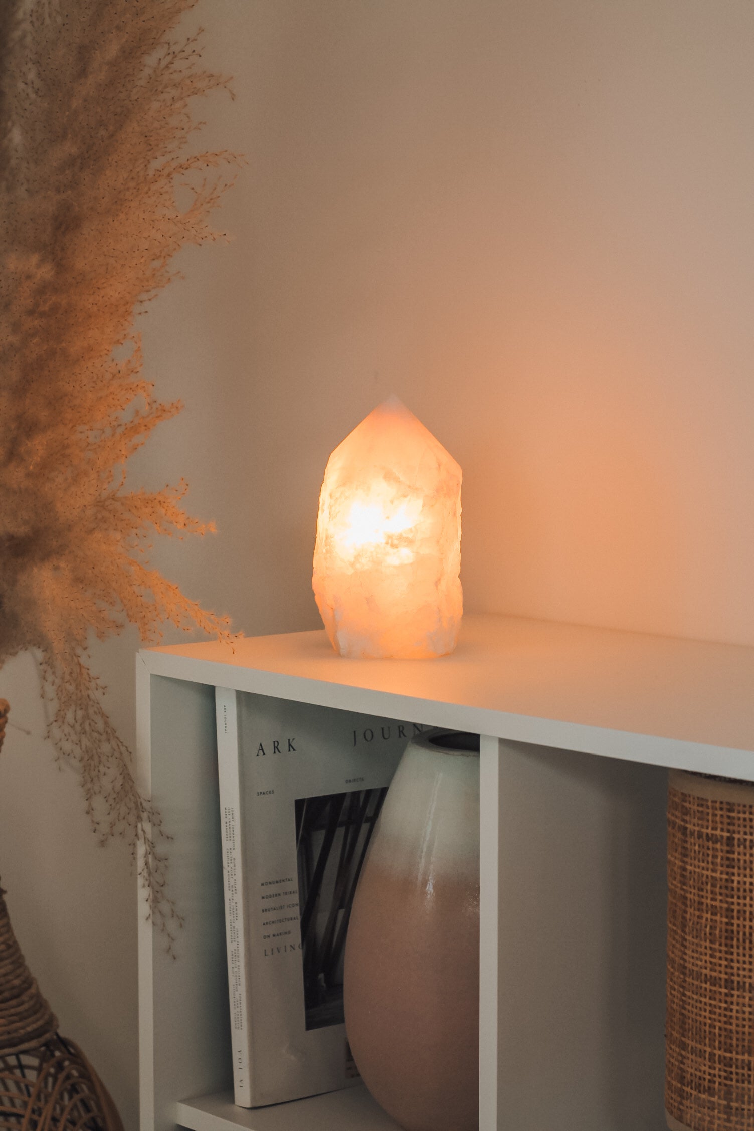 De rozenkwarts punt lamp, brandend op een witte kast. In de kast staat een boek en een grote vaas met witte en roze kleuren. Naast de kast staat nog een stukje van een plant met gedroogde bladeren.