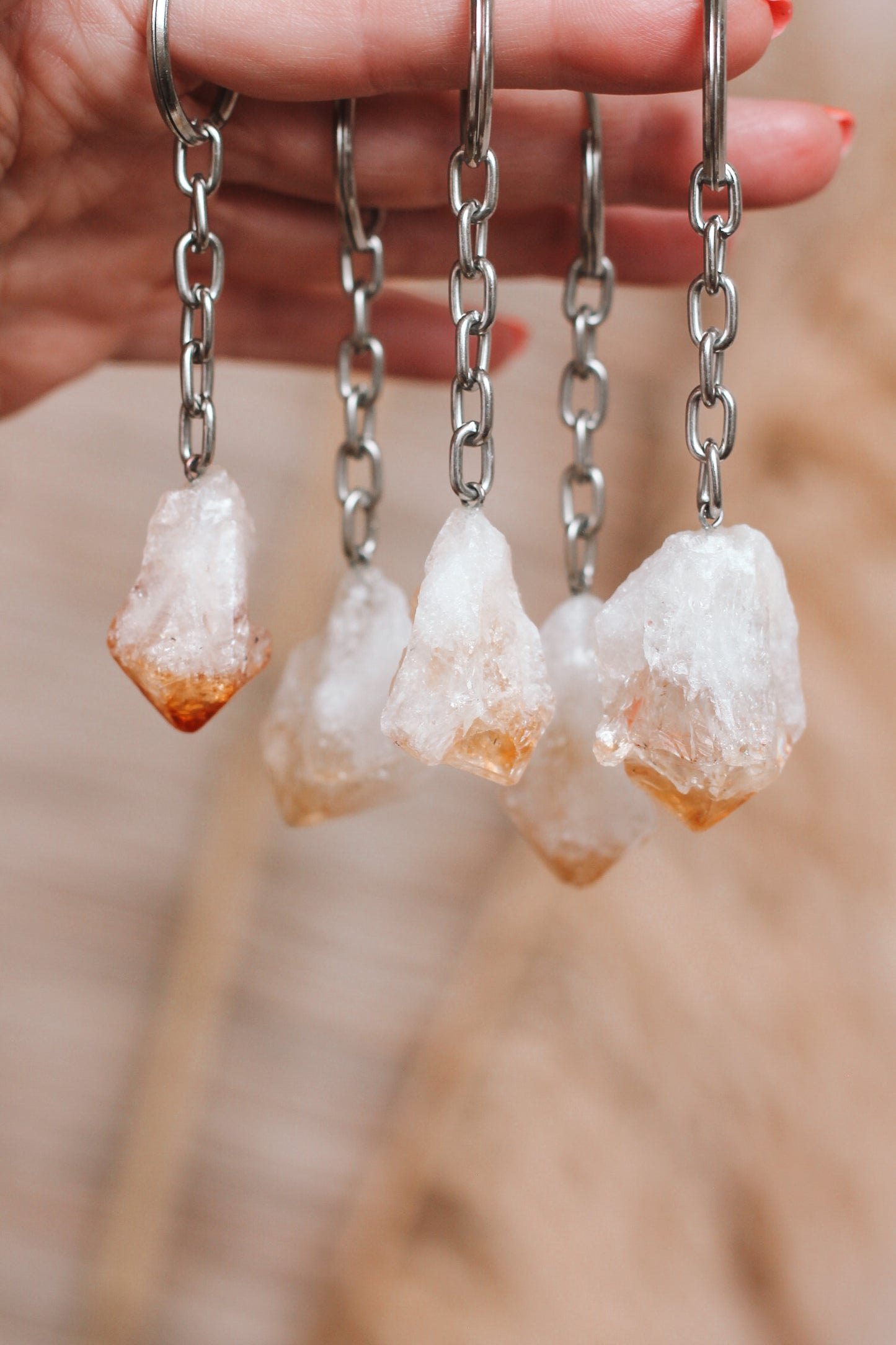 Een hand houdt vier sleutelhangers vast, elk met een stuk citrien kristal. De citrien kristallen zijn ruw en ongepolijst, met een kleurverloop van wit aan de bovenkant naar oranje-bruin aan de onderkant. De sleutelhangers hebben metalen kettingen die de kristallen verbinden met de sleutelhangers.