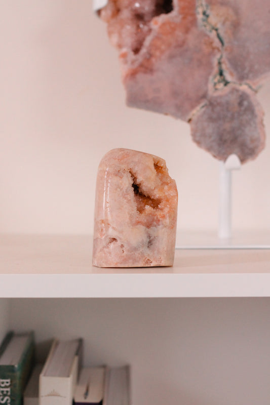 Een roze amethist kristal met een gladde, gepolijste buitenkant en een ruwe, natuurlijke binnenkant. Het kristal staat op een witte plank met enkele boeken eronder en een grotere steen op de achtergrond.