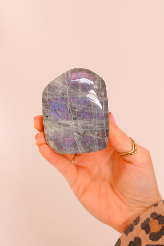 Een hand houdt een roze labradoriet freeform vast. De labradoriet heeft een glanzend oppervlak met iriserende tinten van paars en roze, en vertoont lijnen en patronen die door de steen lopen. De achtergrond is effen en licht van kleur, waardoor de steen goed opvalt.