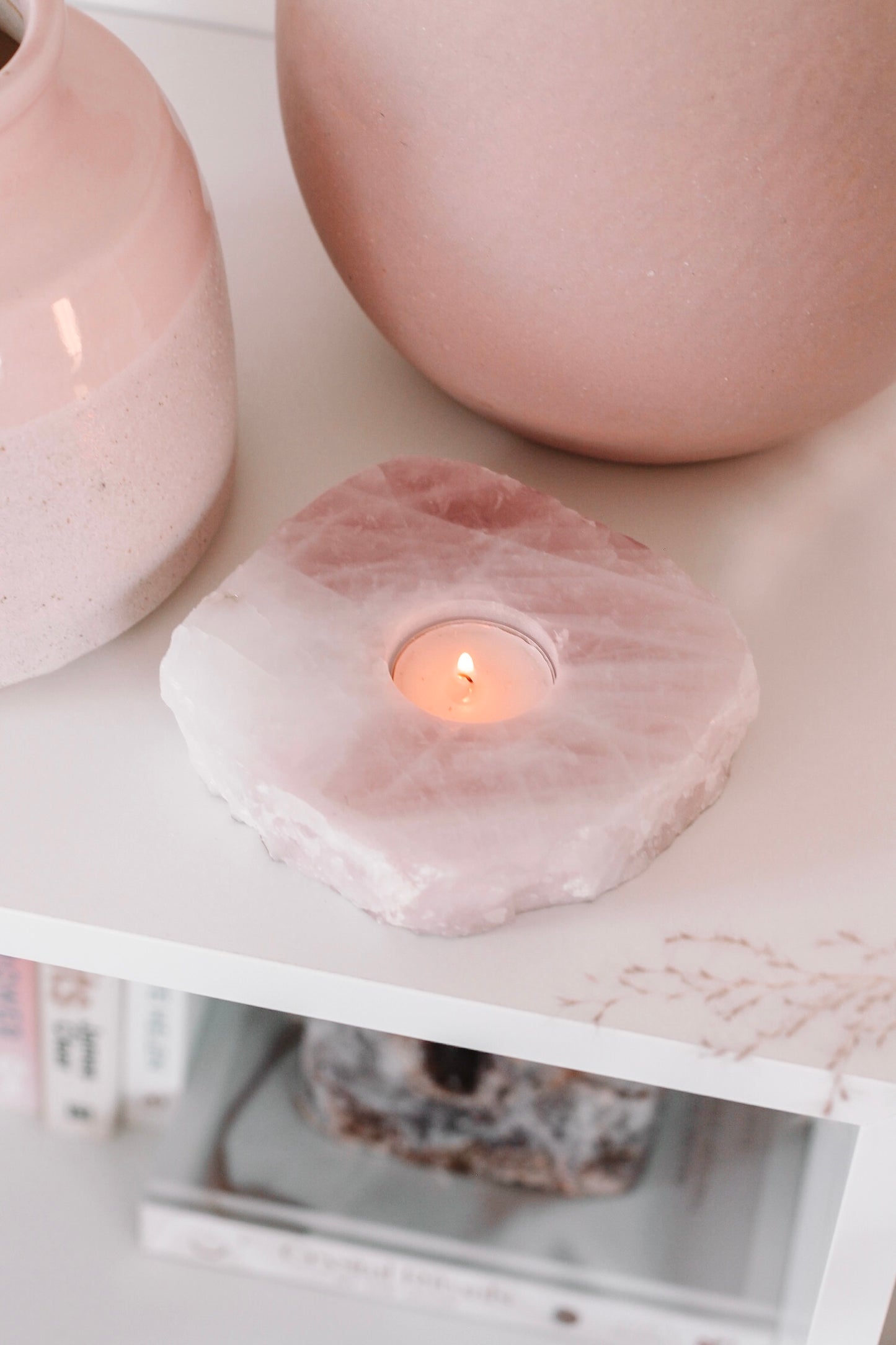 Een brandende theelicht in een houder van rozenkwarts, geplaatst op een witte plank naast twee roze keramische vazen.
