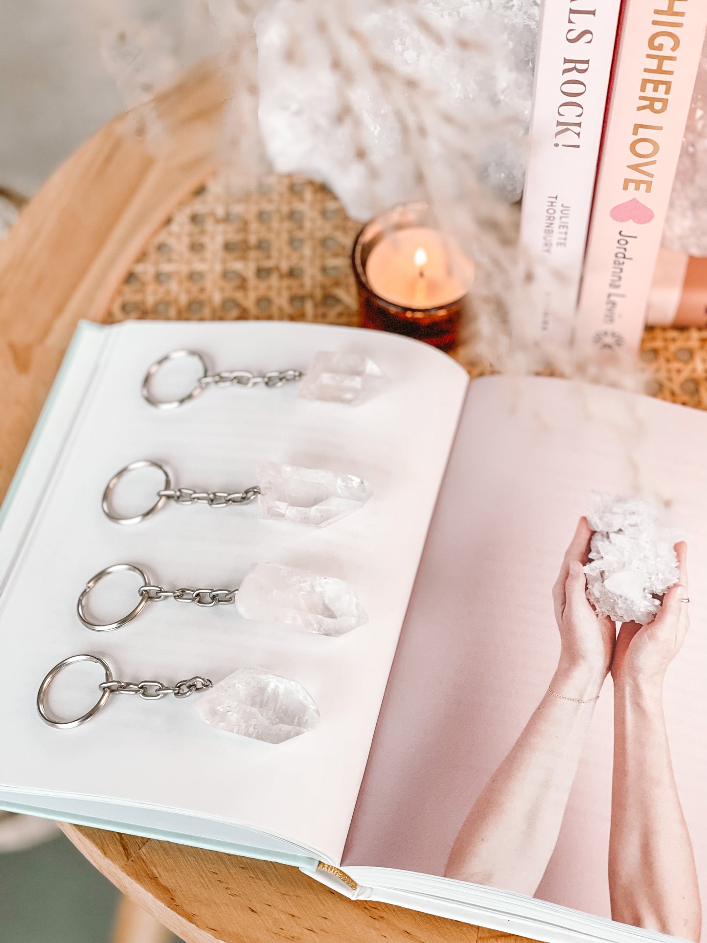 Vier sleutelhangers, elk met een bergkristal eraan bevestigd, liggen op een open boek. De kristallen zijn onregelmatig van vorm en doorschijnend. De sleutelhangers hebben metalen ringen en kettingen die aan de kristallen vastzitten. Op de achtergrond is een brandende kaars in een koperen houder te zien.