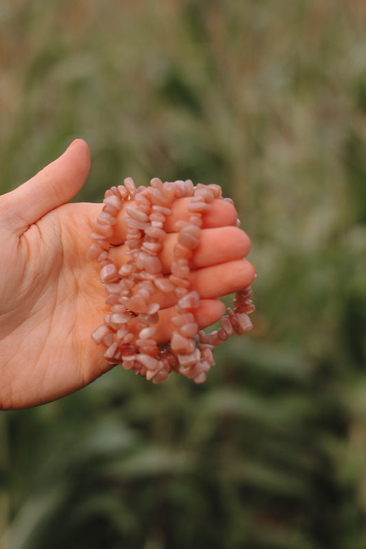 Een hand houdt armbandjes van peach maansteen vast, bestaande uit meerdere kleine, onregelmatig gevormde  maansteen kralen.