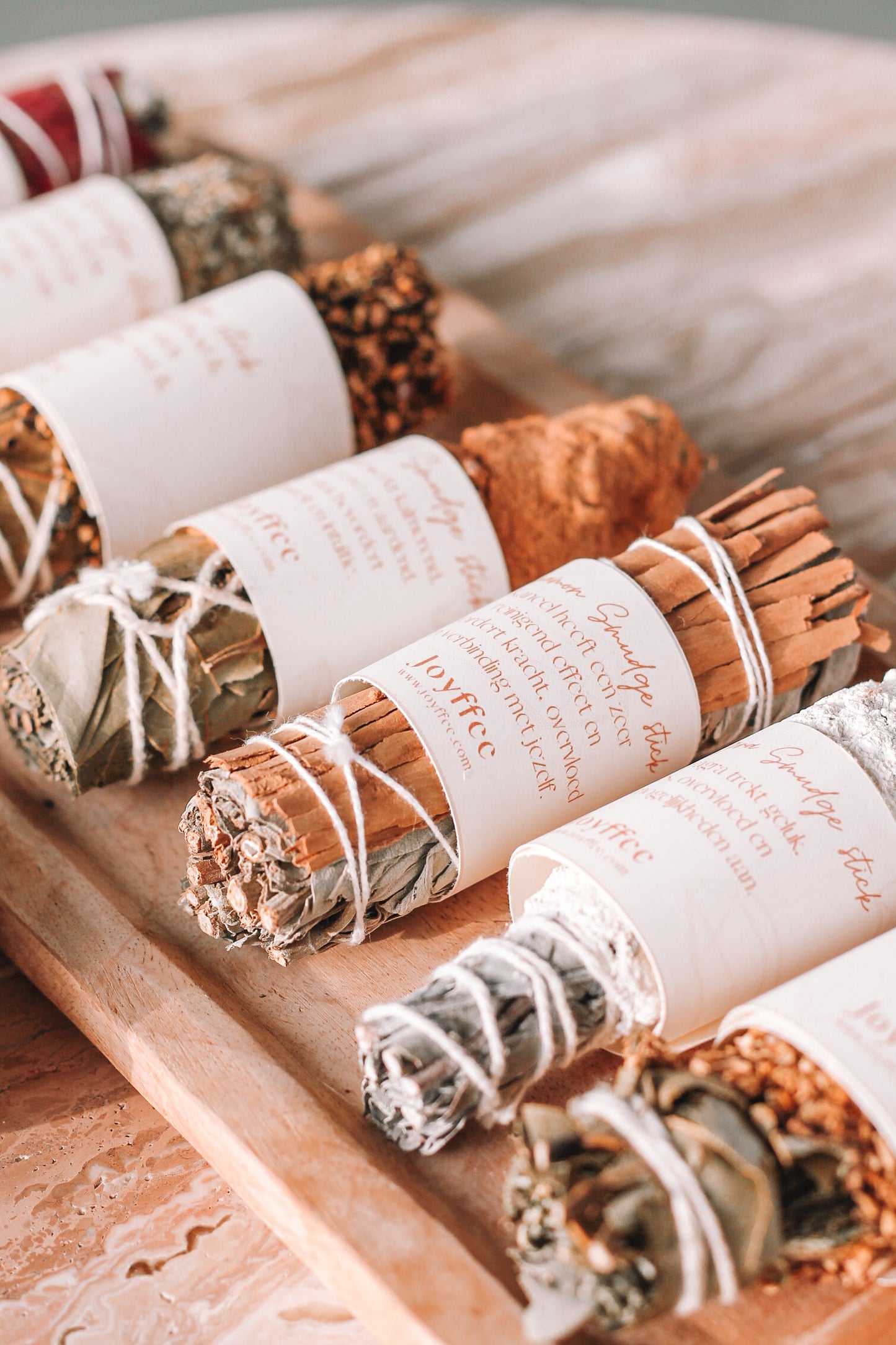 Een houten dienblad met verschillende smudge sticks, elk omwikkeld met wit touw en voorzien van een papieren label. De smudge sticks bestaan uit diverse kruiden en planten.