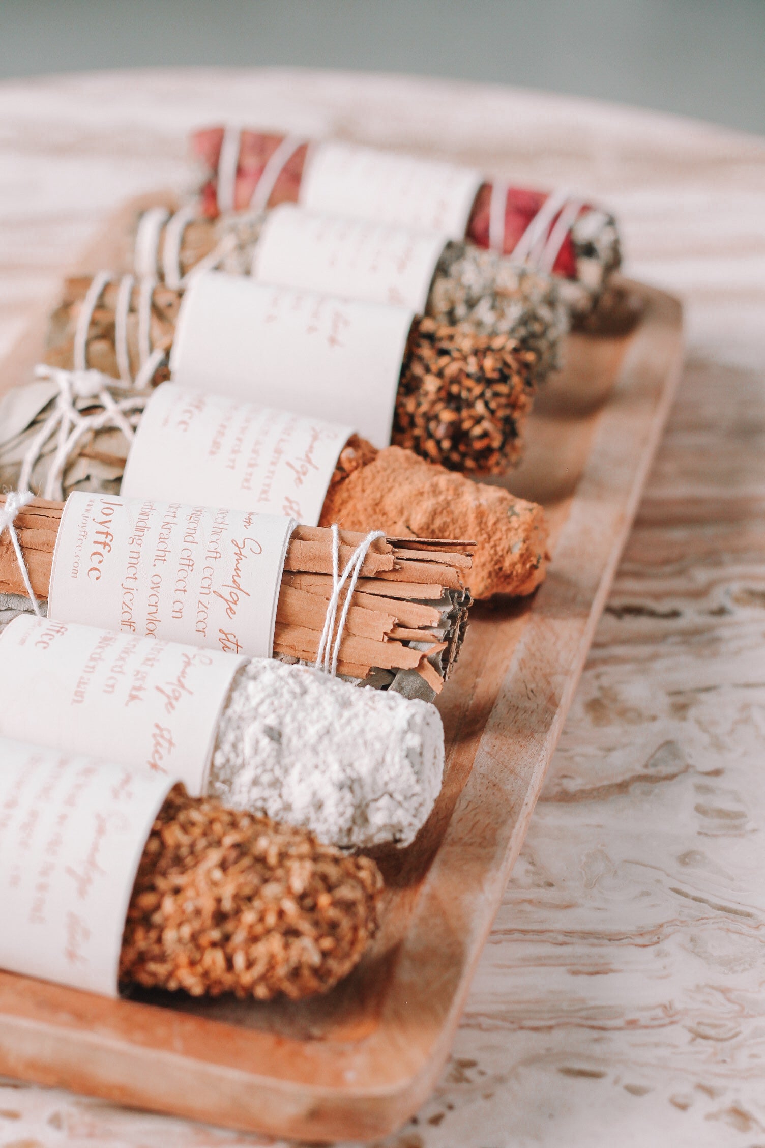 Een houten dienblad met verschillende smudge sticks, elk omwikkeld met wit touw en voorzien van een papieren label. De smudge sticks bestaan uit diverse kruiden en planten.