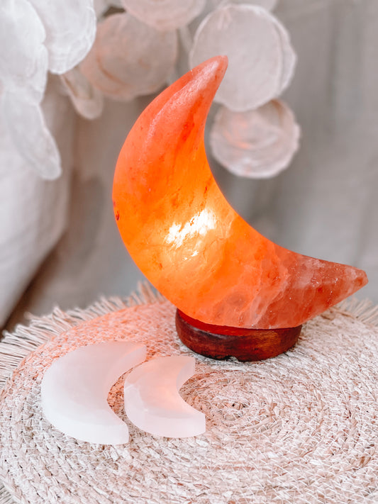 Een  zoutlamp in de vorm van een halve maan, die een zachte, warme gloed uitstraalt. De lamp staat op een ronde, textuurvolle ondergrond met twee kleinere halve maantjes ernaast. 