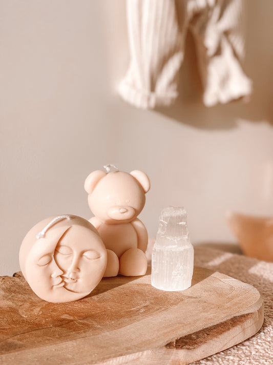 Een seleniet toren geplaatst op een houten oppervlak naast twee kaarsen, waarvan 1 in de vorm van een beertje en de andere in de vorm van 2 halve manen met gezicht.